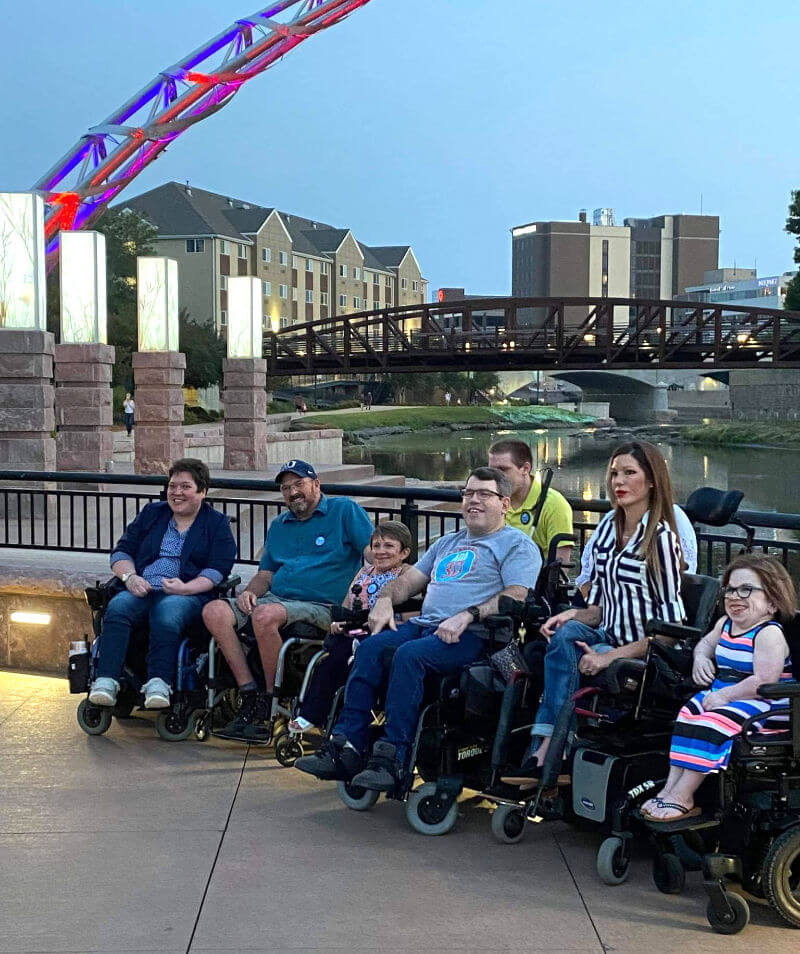 Several people in wheelchairs posing at night underneath the Arch of Dreams statue in Sioux Falls.