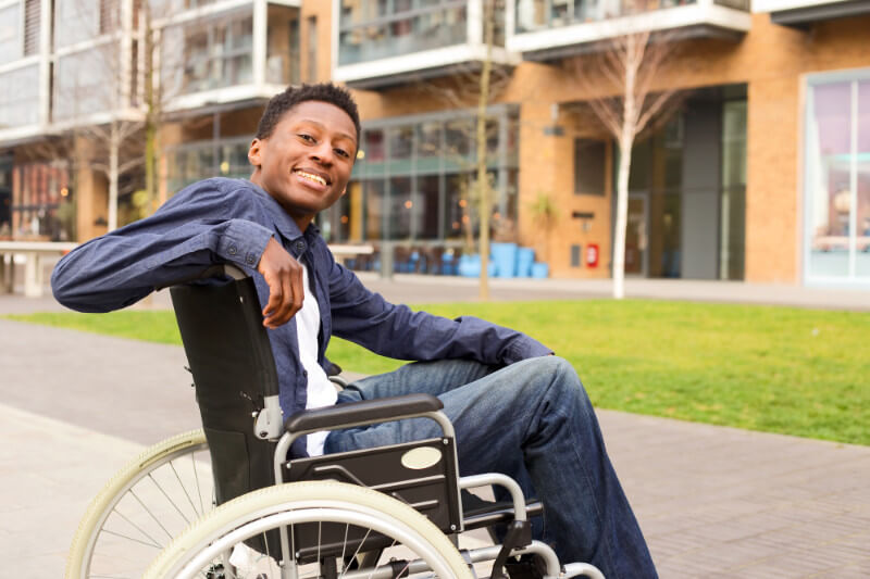 A young African American man smiling and looking back while sitting in his wheelchair outside a college.