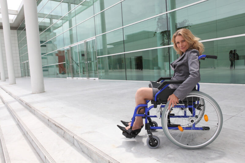A blonde mature woman in a business suit sitting in a wheelchair on the steps outside an office building.