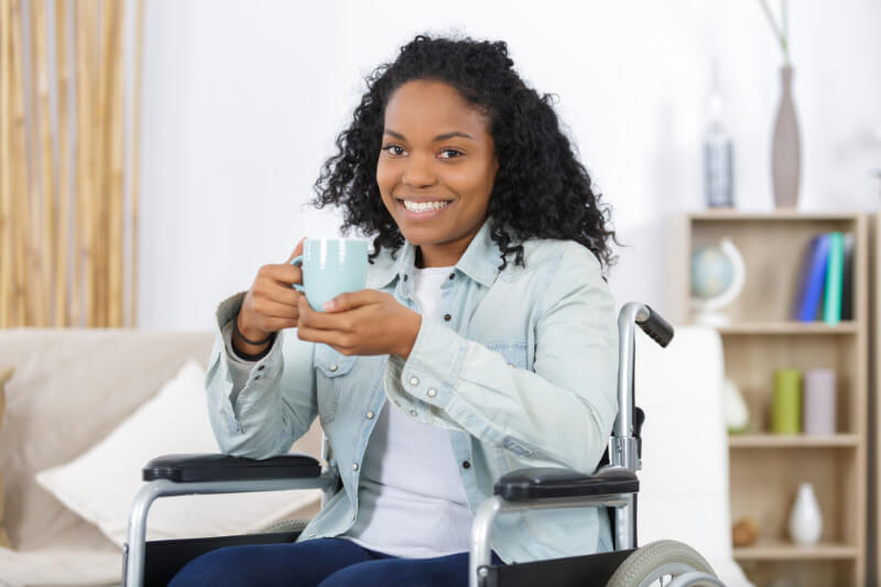 Young disabled African American woman sitting in a wheelchair in an office and smiling and drinking coffee.
