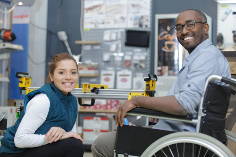 A young caucasian woman kneeling next to a disabled African American man in a wheelchair working in a hardware store.